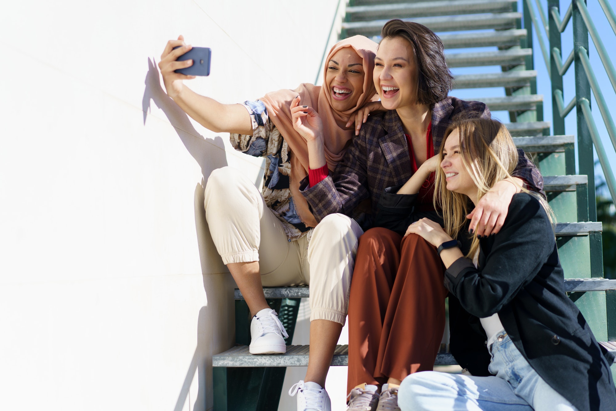Happy multiethnic female best friends taking selfie sitting on stairs in